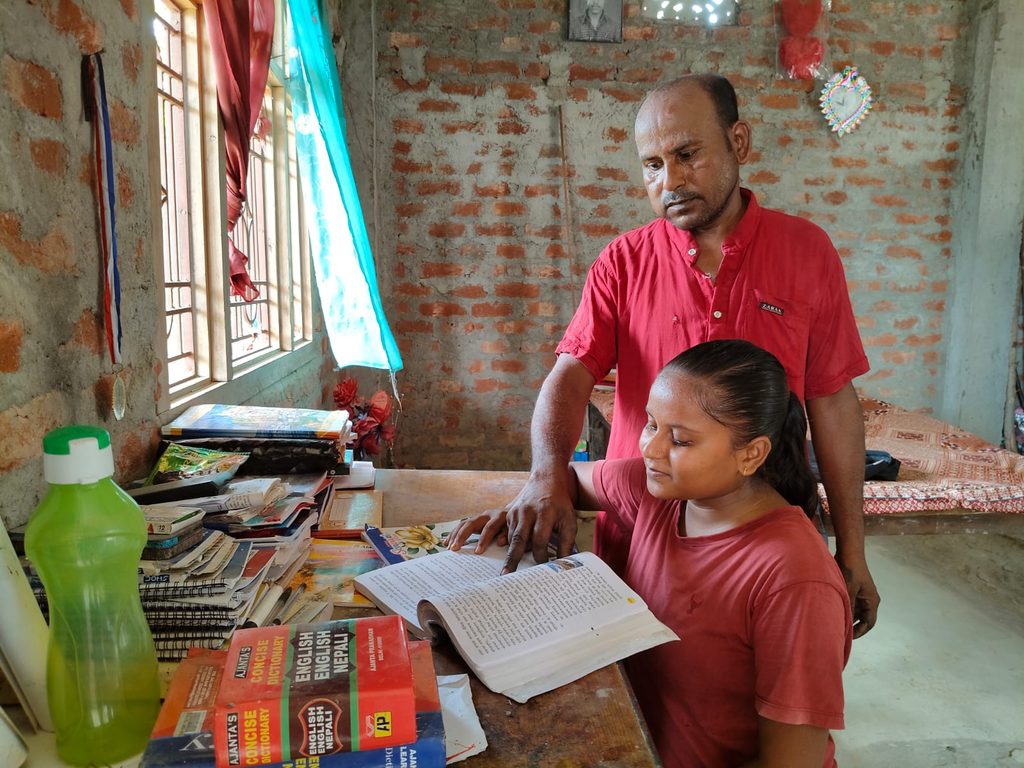 A father supporting his daughter as they read a book together at her study table.
