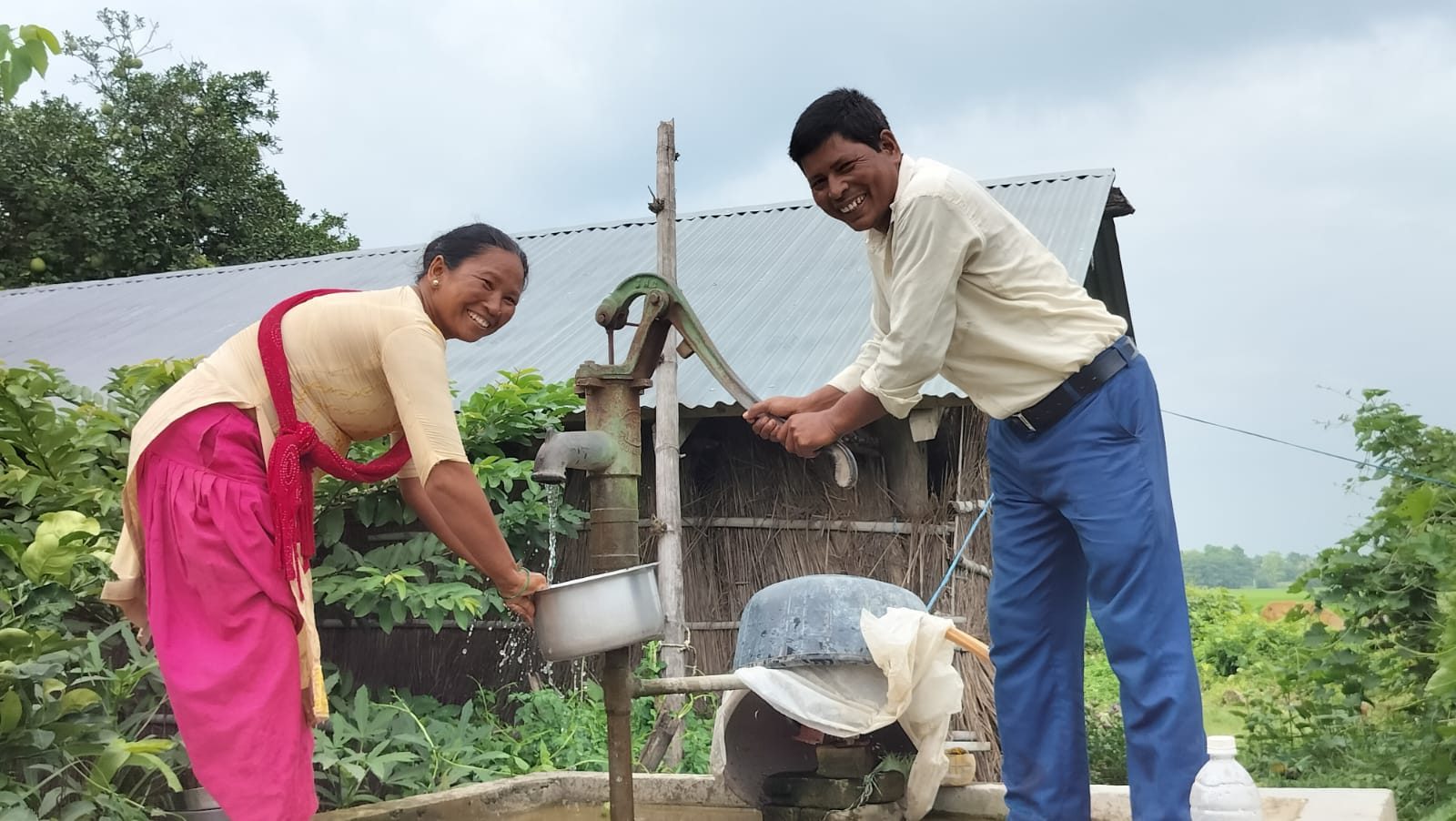 A man in a shirt and trousers is pumping water while a woman washes the dishes together.