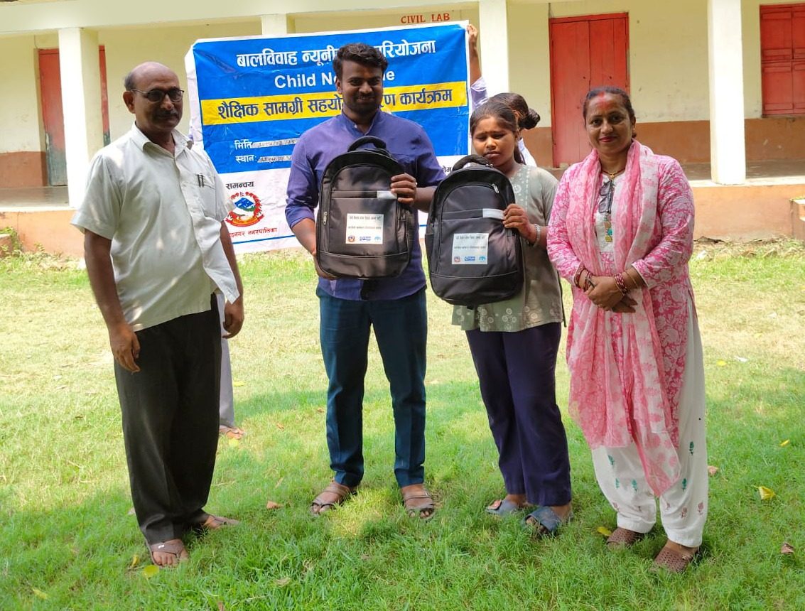 Four people standing in front of banner, two people carrying school bag in their hands.
