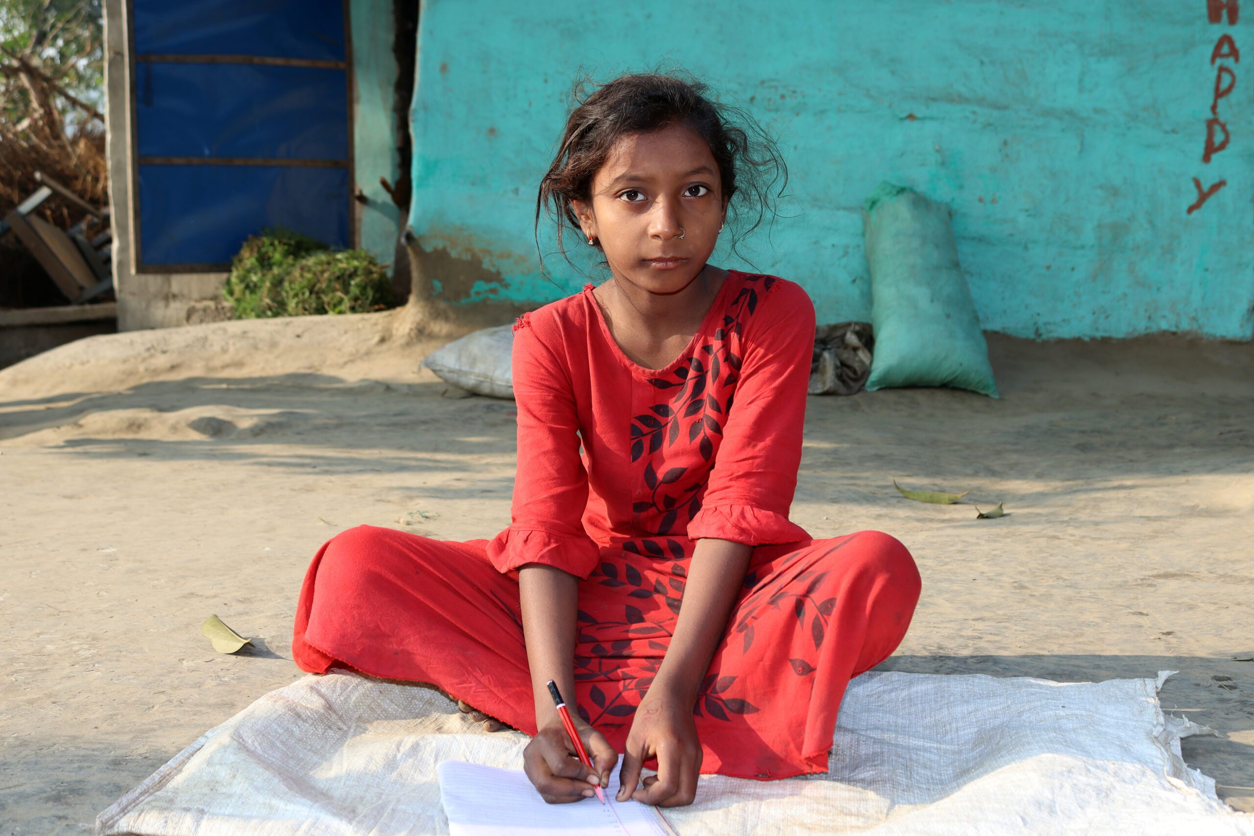 A girl in red dress holding pen and notebook, sitting on the floor in front of her house.