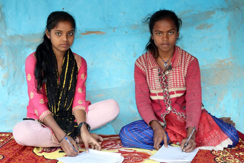 Two girls sitting on a carpet and studying with their pen and notebook.