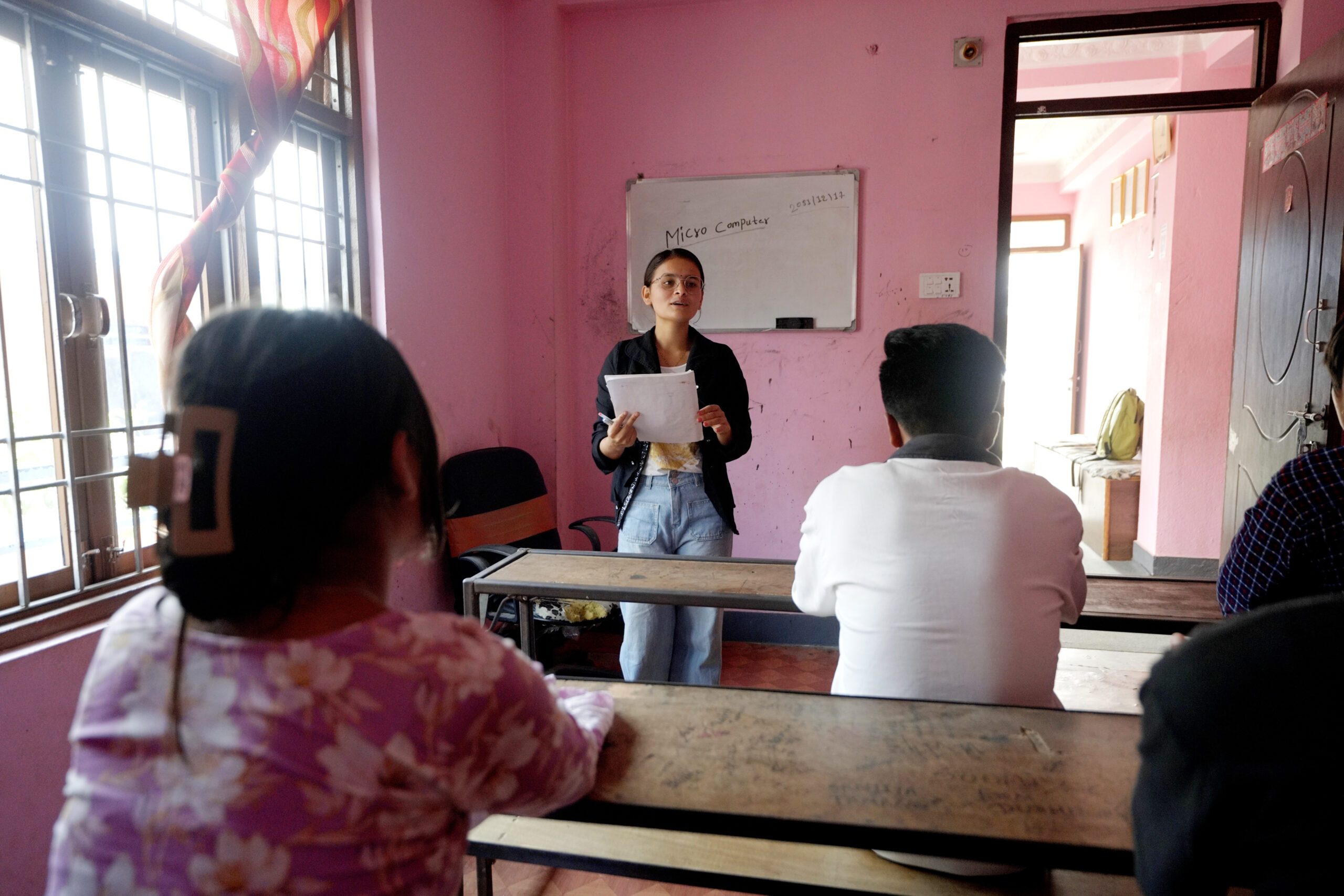 A young woman in black jacket standing in front of white board and holding a note book with other young people sitting and listening to her.