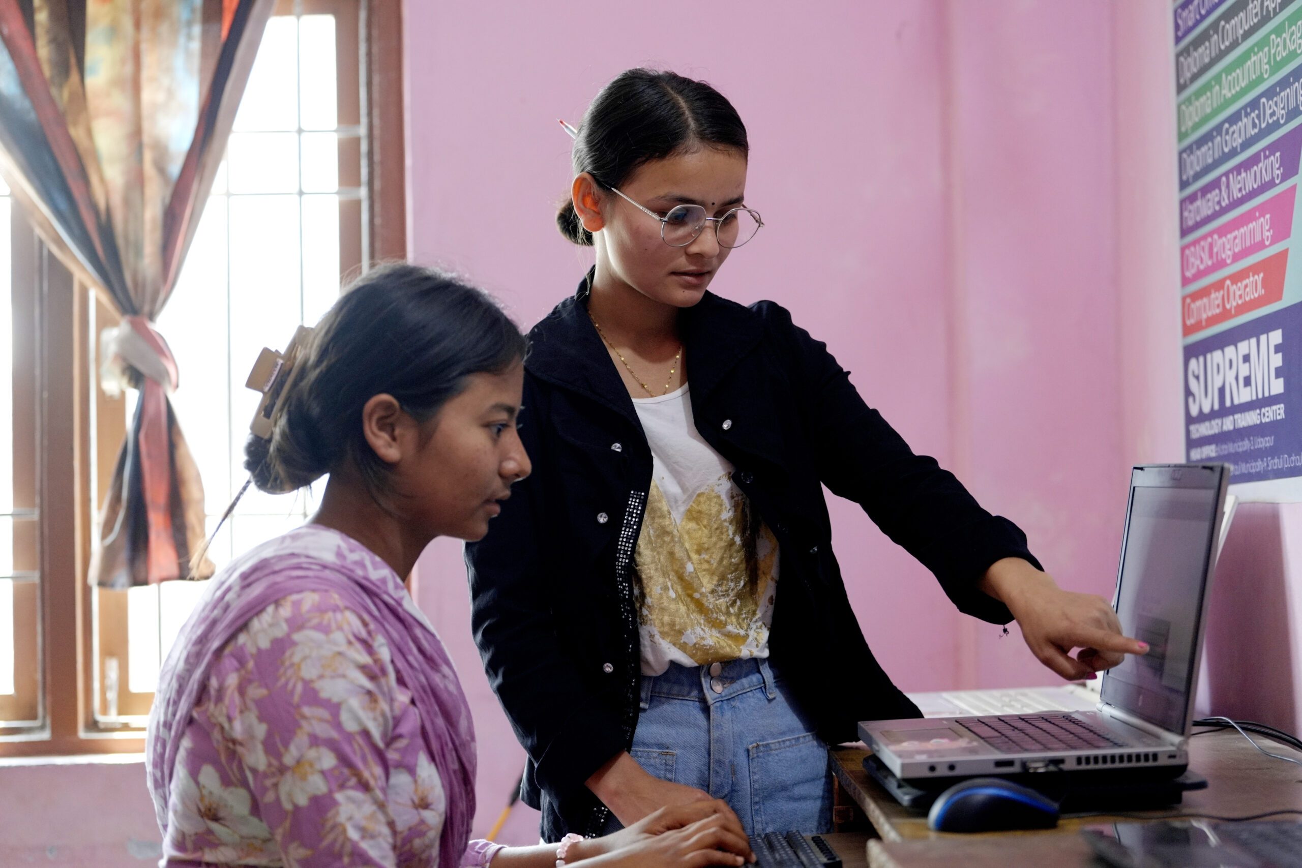 A young women wearing glasses and black jacket pointing in the laptop and a girl sitting and typing using keyboard.