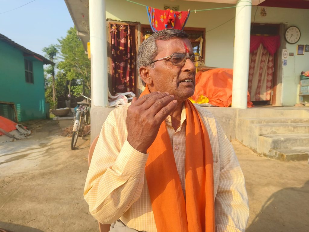 A mid-age man wearing orange shawl, raising his right hand to speak in front of his home.