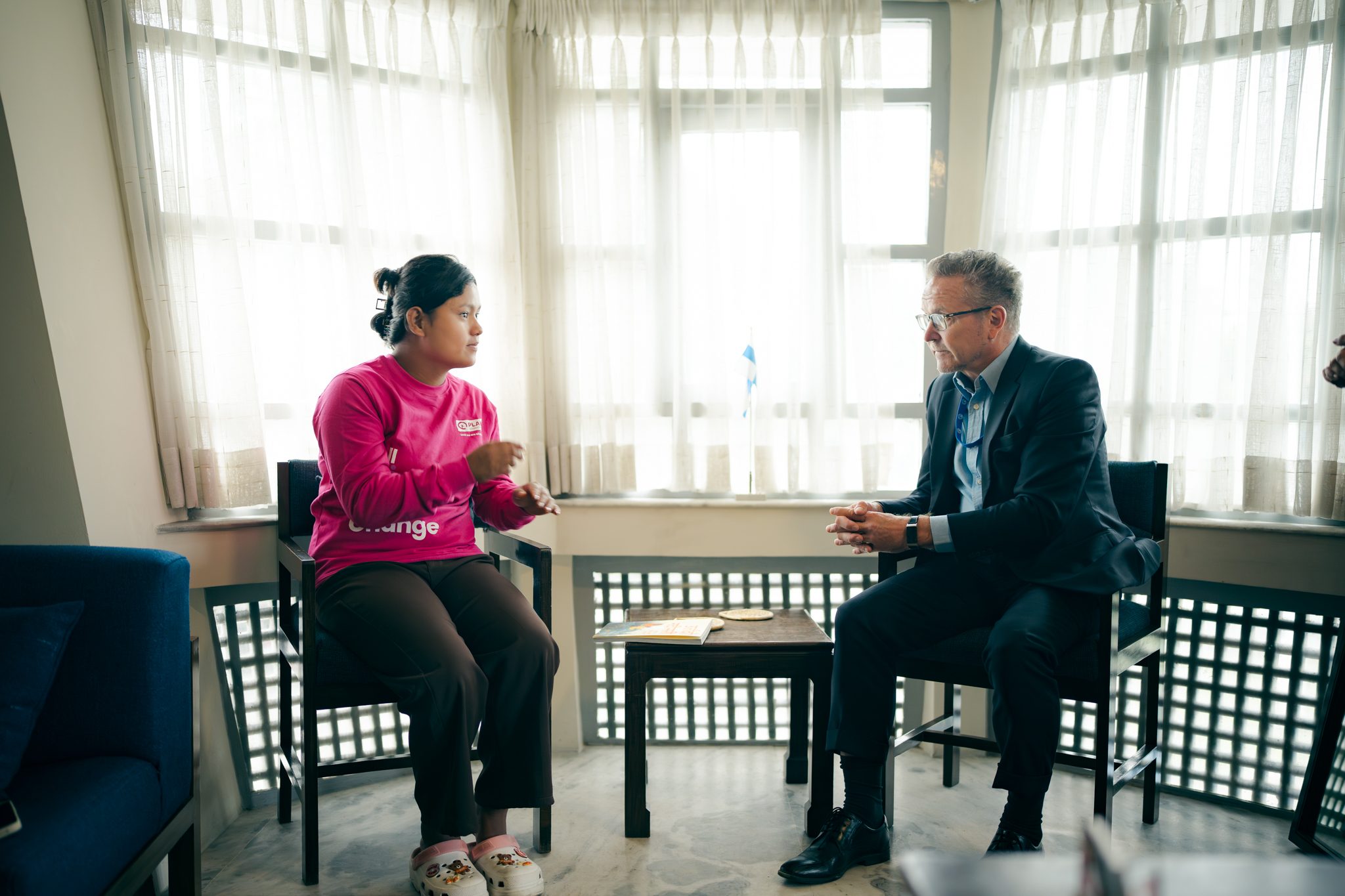 A girls in pink (left) and a man on suite (right) sitting on chairs facing to each other for conversations.