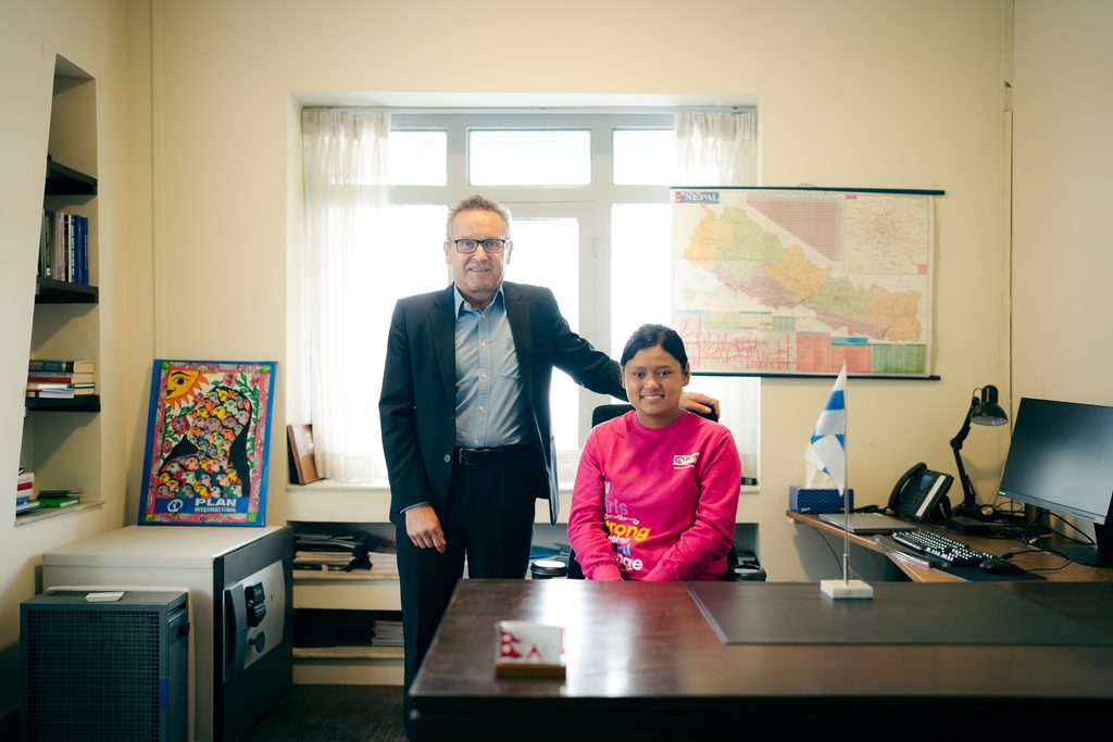 A man wearing black suite standing beside a girl wearing pink hoddie siting on a chair.