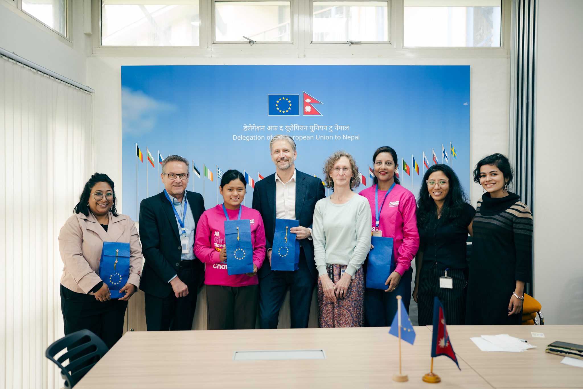 8 people together posing for a photo with flag of EU and Nepal beside.