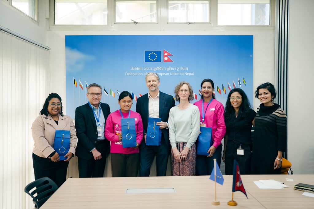 8 people together posing for a photo with flag of EU and Nepal beside.