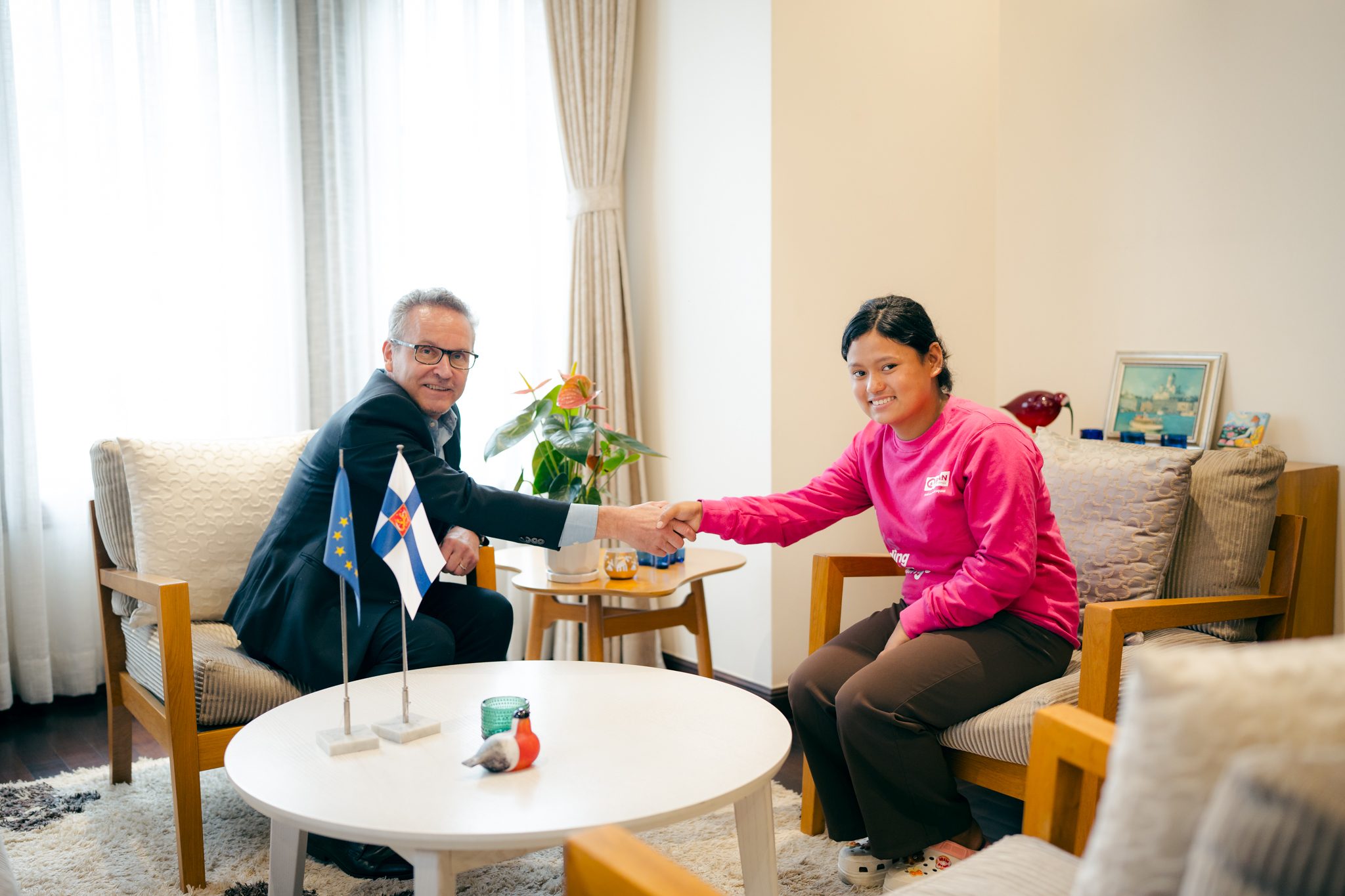 A man in black suit on left and girl in pink in right shaking their hands.