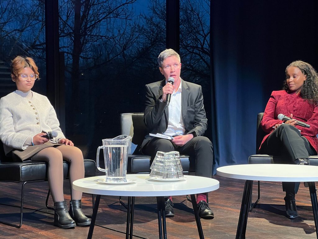 Three panellists sit on chairs with microphones on small tables in front of them: Smarika in an off-white coat on the left, Julian in a black suit in the middle, and Aldine in red on the right.