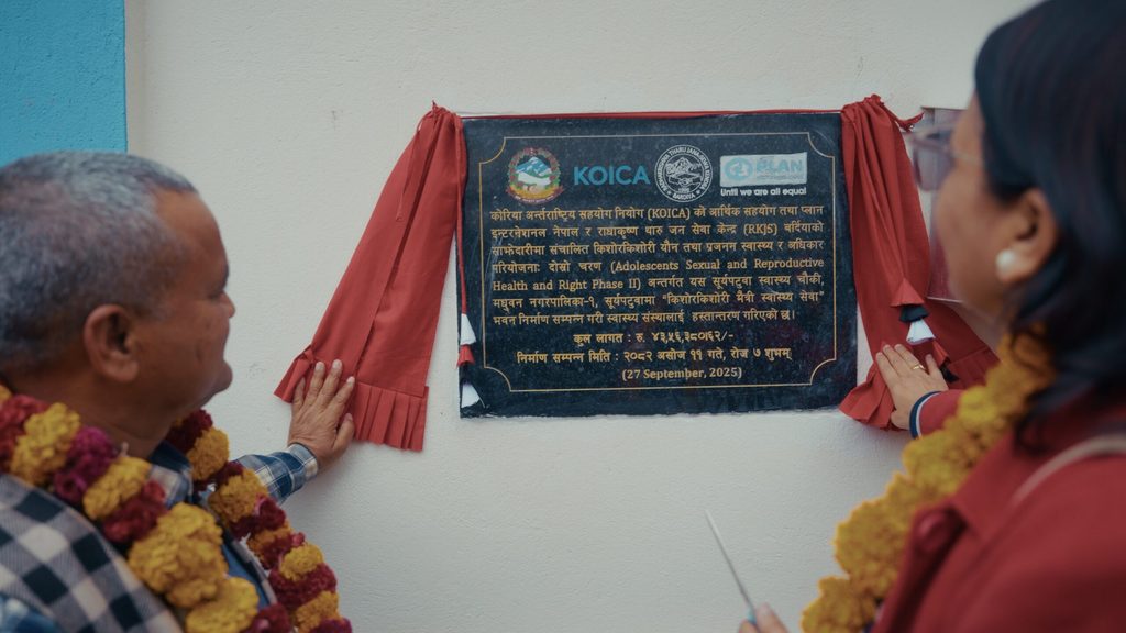 A man on the left and a woman on the right look at a plaque displaying the logos of the Government, KOICA, RKJS, and Plan International.