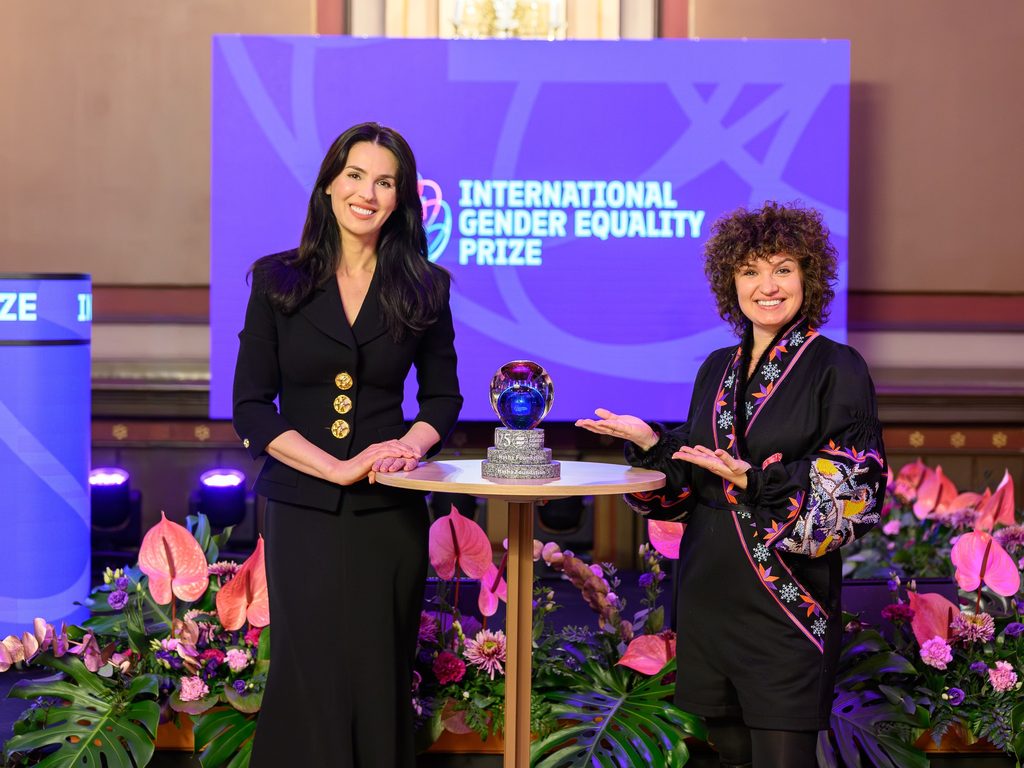 Two women in black dresses stand on a stage receiving the International Gender Equality Prize award.