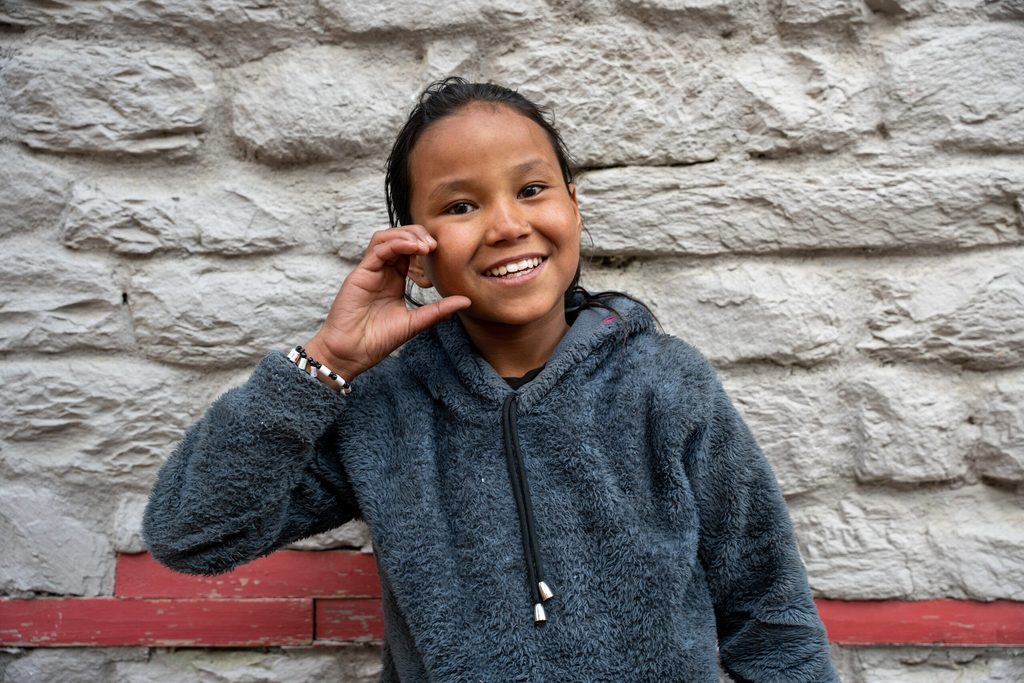 A young girl wearing black poses in front of a wall, making a “C” shape on her face with her hand.