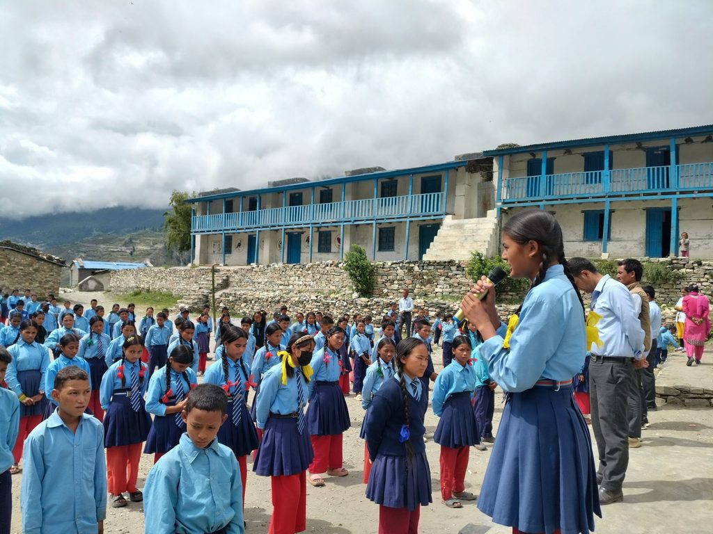 Ambika, wearing her school uniform, speaks into a microphone while standing in front of other students during a school assembly.