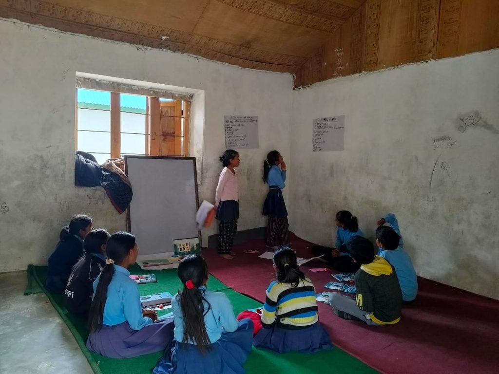 A room with window and white board where a group go girls are siting in circle and watching two girls who are standing and looking into the chart paper pasted on the wall.