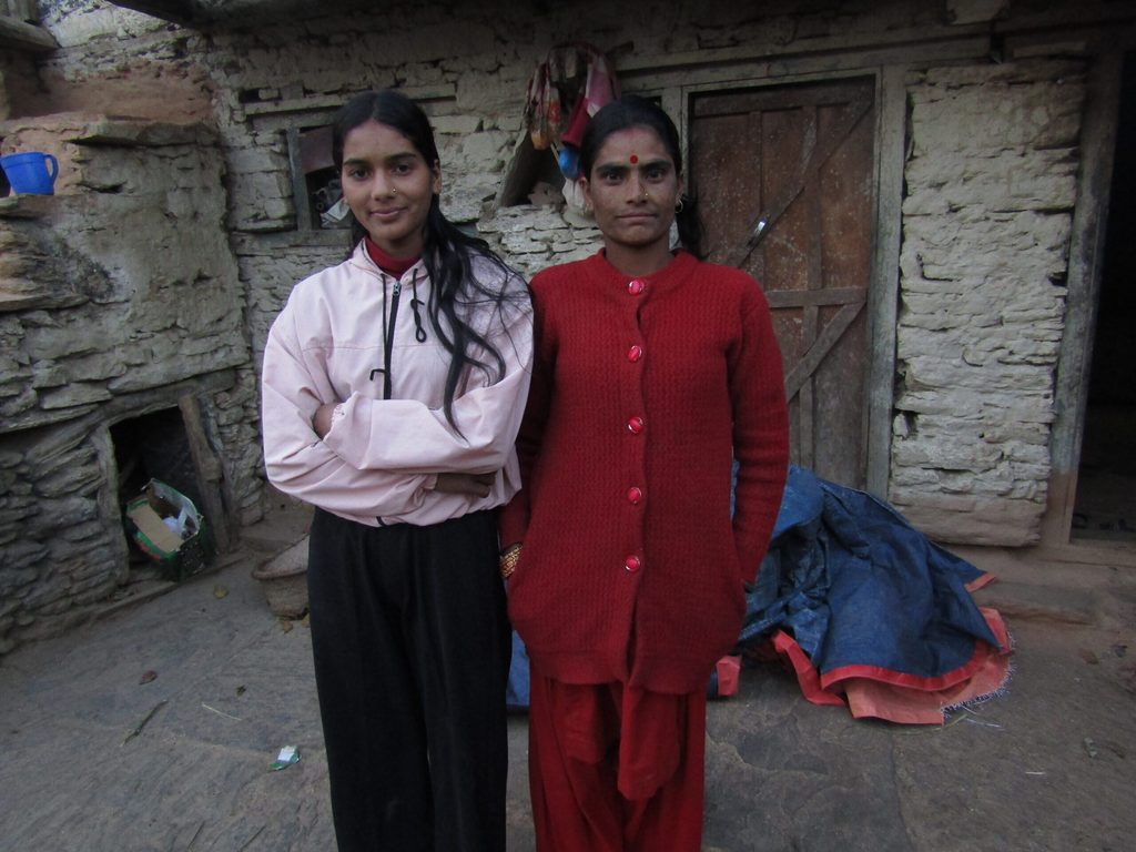 Niruta in a light pink hoodie stands next to a woman in a red sweater in front of their house.