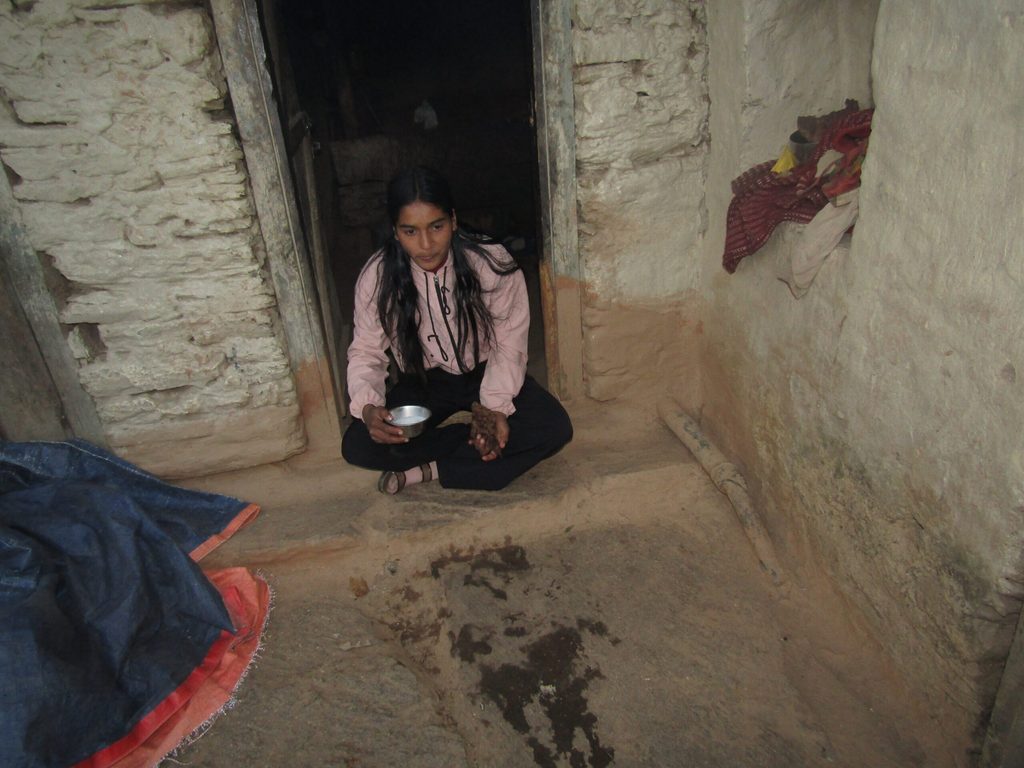 Niruta in a light pink hoodie sits in a doorway, holding a bowl of milk.