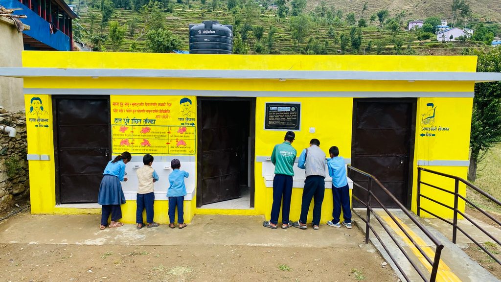 A group of students wash their hands in front of a yellow-painted toilet with ramps.