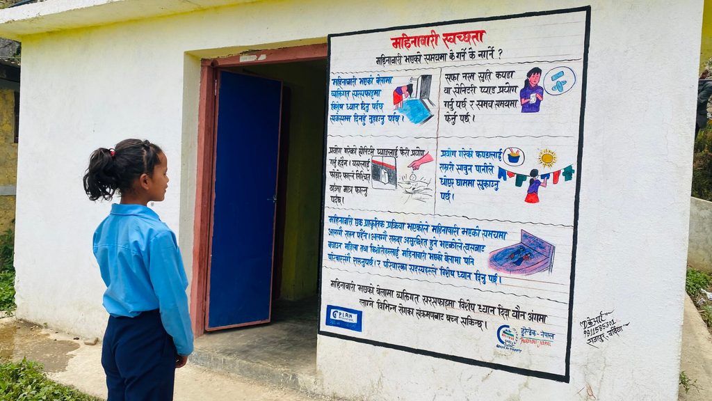 A female student in a light blue school shirt and dark blue trousers looks at a wall painted with information about menstrual health and hygiene.