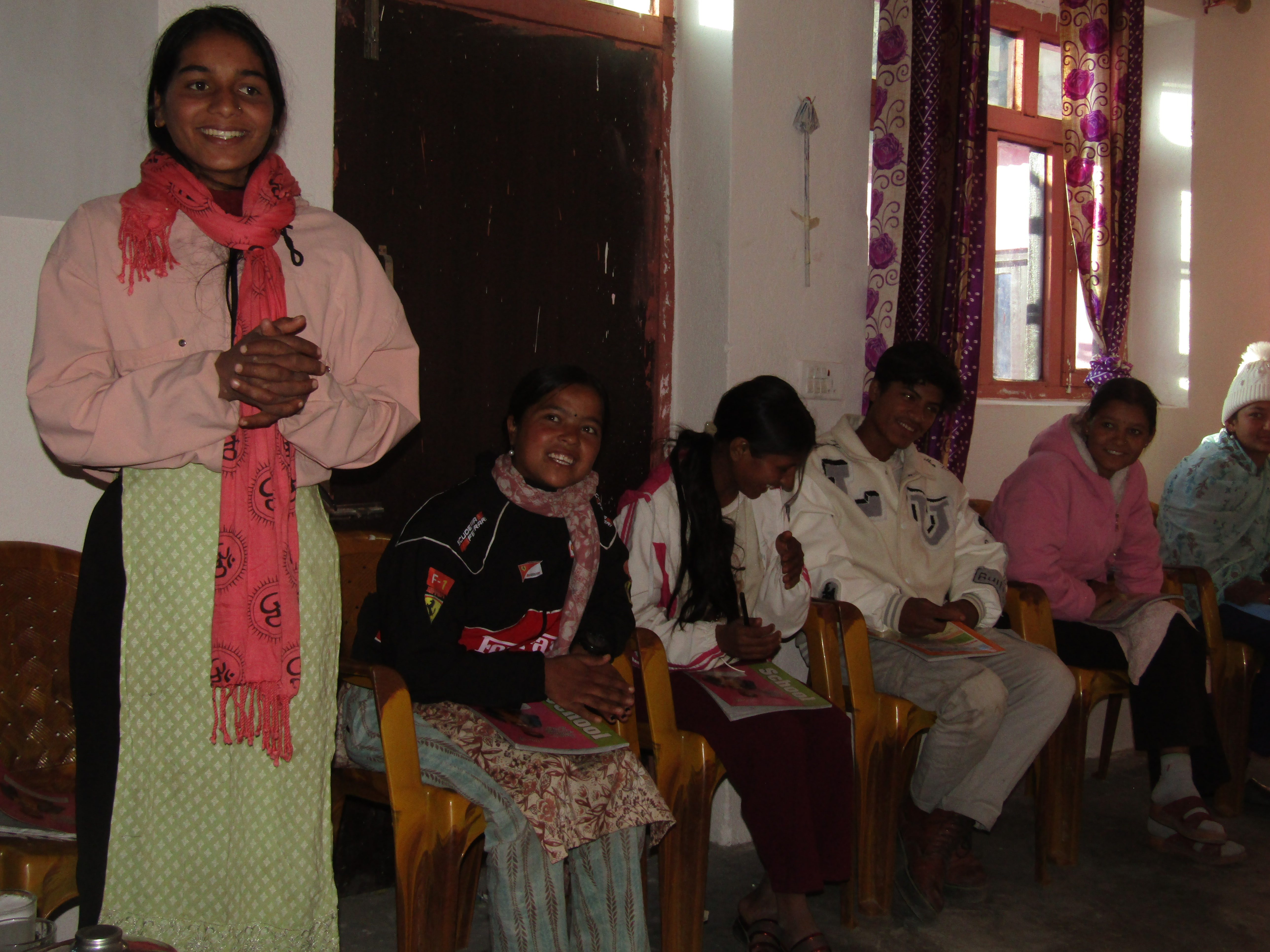 Niruta in a light pink hoodie stands and smiles while other youths sit on chairs holding notebooks.