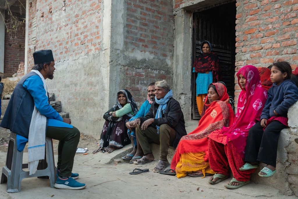 Sadre wearing blue shirt and black cap siting on a chair and talking to a group of community people.