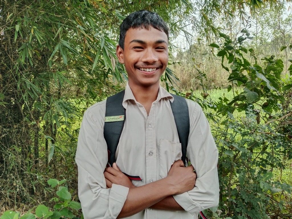 Rohit in his school uniform carrying bag pack, smiling and standing in front of bushes.