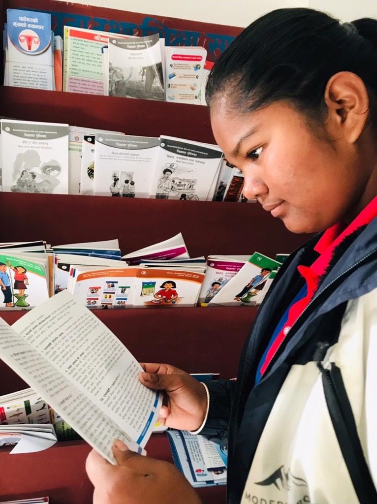 Mira is standing and holding informative book on adolescent SRHR and reading it. 