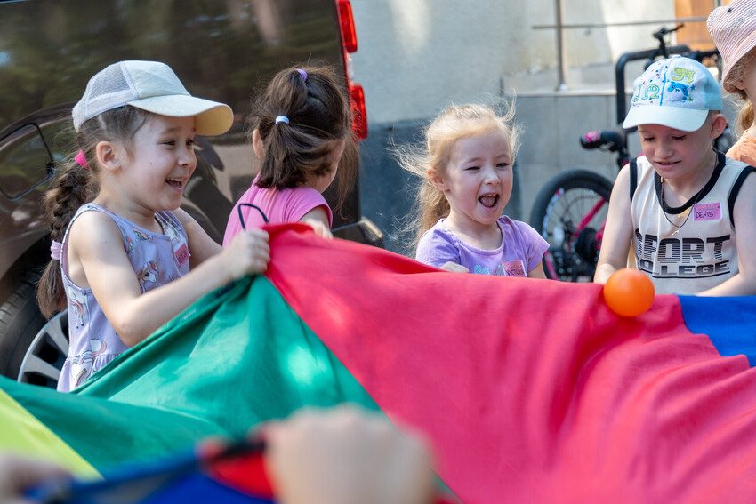 Children are lifting the edge of a toy parachute and laughing. 